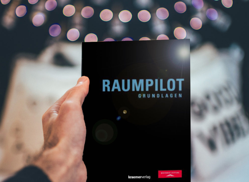 Eine Person hält ein Buch mit dem Wort „Raumplot“ hoch, das bei der Wüstenrot Stiftung zum kostenlosen Download erhältlich ist.